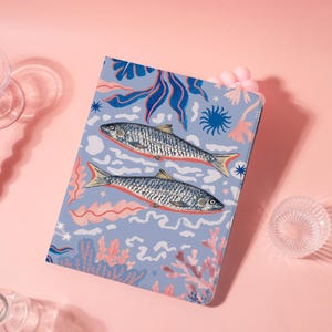 Könnte beinhalten: Ein Notizbuch mit hellblauem Einband, das zwei Fischillustrationen zeigt. Die Fische sind silbern mit orangefarbenen Akzenten, vor einem Hintergrund aus Korallen, Seetang und sternförmigen Designs in Rosa und Blau.