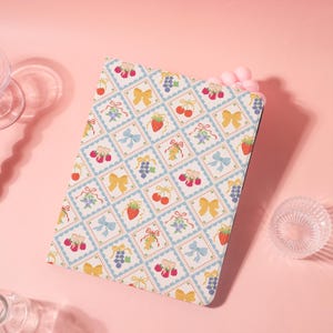 May include: A patterned tablet case featuring a repeating design of fruits, bows, and floral elements in a grid pattern. The design includes strawberries, cherries, grapes, and bows in a pastel color palette of blue, yellow, and red on a cream background.