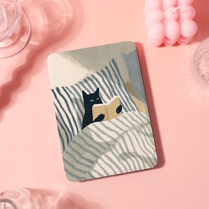 May include: A rectangular art print featuring a black cat reading a book in bed. The cat is under a striped blanket, with a neutral color palette of gray, beige, and blue. The background is a soft pink.