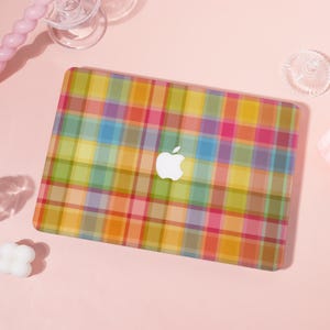 May include: A laptop with a colorful plaid pattern case. The case features a vibrant mix of colors including pink, yellow, green, blue, and orange. A white Apple logo is visible on the case. The laptop is on a pink surface.