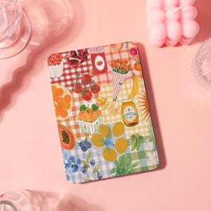May include: A rectangular, colorful placemat with a patchwork design featuring illustrations of fruits, vegetables, and honey. The design includes oranges, tomatoes, blueberries, and a jar of honey. The background is a mix of gingham and floral patterns.