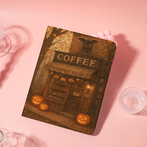 May include: A notebook with a Halloween-themed illustration of a coffee shop. The cover features the word "COFFEE" above the shop's awning, with pumpkins and a bat. The shop has a "BOOKS" sign and an "OPEN" sign. The color palette includes orange, brown, and gold.