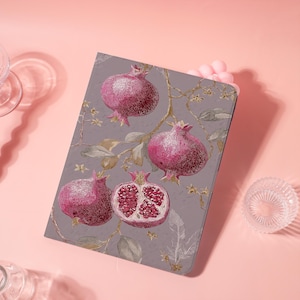 May include: A rectangular notebook featuring a botanical design of pomegranates in shades of pink and red against a gray background. The design includes branches, leaves, and floral accents. One pomegranate is cut open, revealing the seeds.