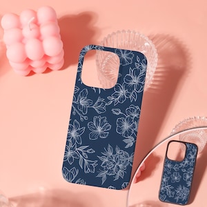 May include: A navy blue phone case with a white floral design. The pattern features detailed outlines of flowers and leaves, offering a delicate and elegant look. The image includes a smaller case, a pink candle, and a glass dish.