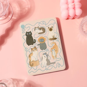 May include: A beige tablet case featuring a playful illustration of various cartoon cats. The cats are in different poses and colors, with a light blue maze-like design around them. A ball of yarn is in the upper left corner.