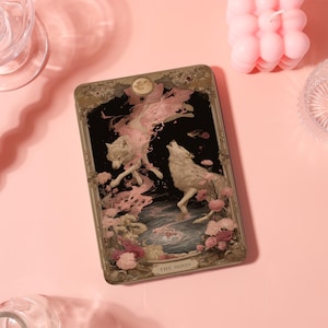 Könnte beinhalten: Eine rechteckige Tarotkarte mit schwarzem Hintergrund, die zwei weiße Wölfe, rosa florale Details und einen Vollmond zeigt. Die Karte hat einen verzierten Goldrand und den Text "THE MOON" unten.