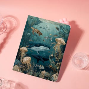 May include: A rectangular notebook with an underwater illustration of whales, jellyfish, and a sea turtle. The artwork uses blue, green, and gold tones, showcasing marine life and plants. The notebook is displayed on a pink surface.