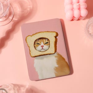 May include: A pink tablet case featuring a whimsical illustration of a cat with a slice of bread around its head. The cat has orange and white fur and a slightly grumpy expression. The bread is golden brown.