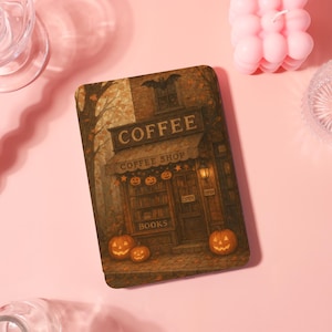 Könnte beinhalten: Ein rechteckiges Notizbuch zum Thema Halloween, das ein Café mit Kürbissen, einer Fledermaus und dem Wort "COFFEE" zeigt. Der Laden hat ein "OPEN"-Schild und das Wort "BOOKS" ist sichtbar. Die Farbpalette umfasst Braun-, Orange- und Gelbtöne.