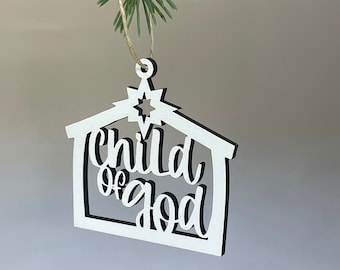 Könnte beinhalten: Ein weiß-schwarzer Weihnachtsbaumschmuck aus Holz mit dem Schriftzug "Child of God". Der Ornament, geformt wie ein Haus mit einem Stern, hängt an einer Schnur an einem grünen Kiefernzweig. Schlichtes und elegantes Design.