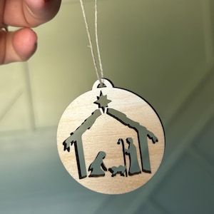 May include: A wooden Christmas ornament with a cutout design of the nativity scene. The ornament is light brown and features a star, a stable, Mary, Joseph, and the baby Jesus.