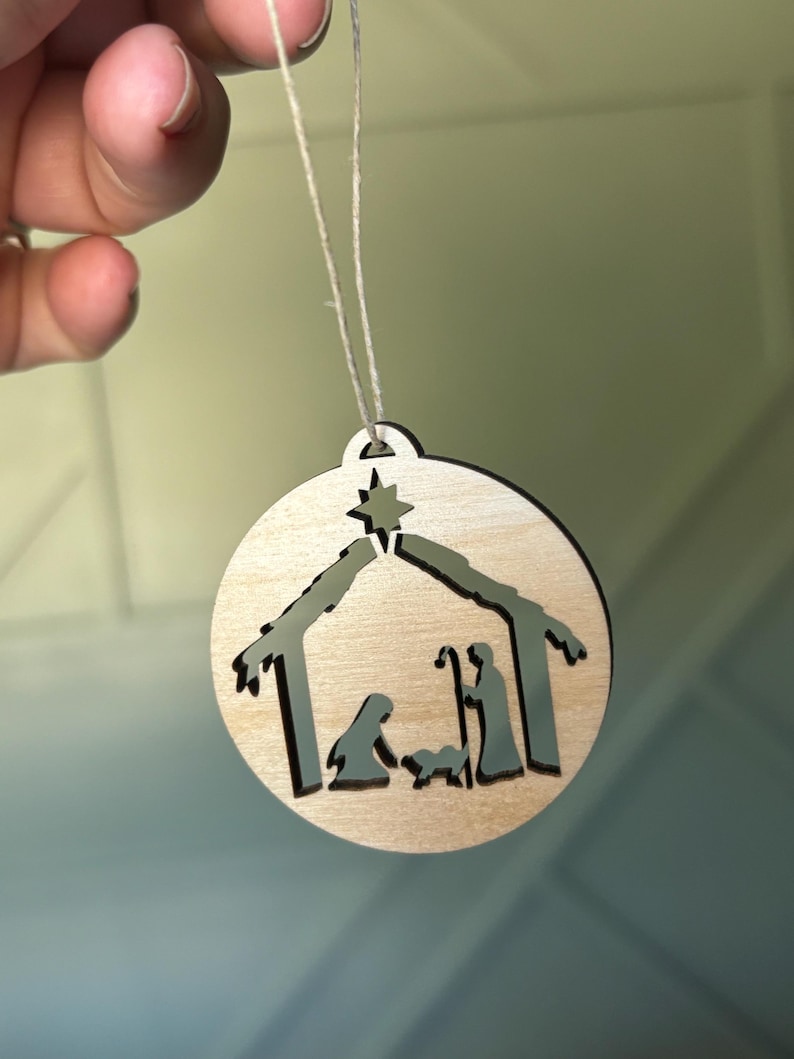May include: A wooden Christmas ornament with a cutout design of the nativity scene. The ornament is light brown and features a star, a stable, Mary, Joseph, and the baby Jesus.