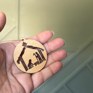 May include: A wooden Christmas ornament featuring a cutout design of the nativity scene. The ornament is round and has a brown wood finish. A star is cut out at the top of the ornament.