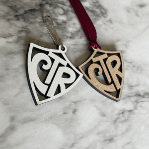 May include: Two wooden ornaments shaped like shields with the letters "CTR" cut out in the center. One ornament is white with black lettering and the other is brown with black lettering. A red ribbon is attached to the brown ornament.