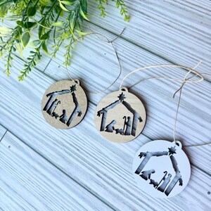 May include: Three wooden Christmas ornaments with a cutout design of the nativity scene. The ornaments are in natural wood, stained wood, and white.