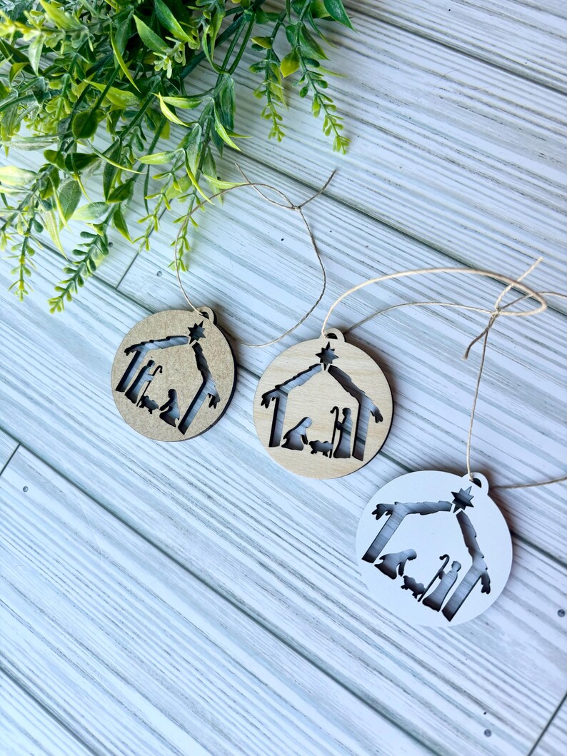 May include: Three wooden Christmas ornaments with a cutout design of the nativity scene. The ornaments are in natural wood, stained wood, and white.