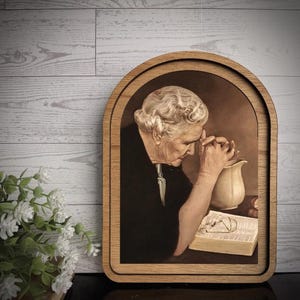 May include: A framed arched wall art piece featuring a sepia-toned illustration of an elderly person with wavy hair, wearing a dark garment, with hands clasped in prayer beside an open book and a ceramic pitcher on a table surface.