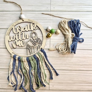 May include: A wooden circular wall hanging with the words "Walk with Me" and an image of Jesus. The hanging has a macrame fringe in blue, green, and cream colors. Additional cream and blue macrame rope and wooden beads are included.