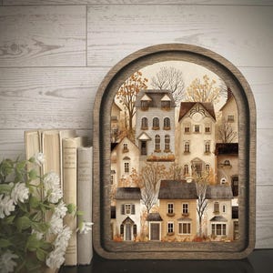 May include: An arched wooden frame containing a multi-layered paper art scene of a village with houses in shades of cream, beige, and brown. The scene features bare trees and autumn foliage, with small pumpkins placed near the building entrances.