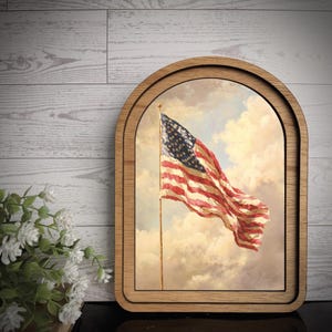 May include: A framed print of the American flag waving in the wind against a cloudy sky. The print is set within a wooden, arched frame. The flag features red and white stripes and a blue field with white stars.