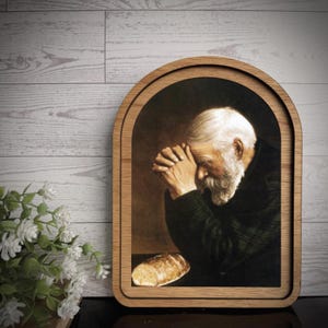 May include: An arched wooden frame containing a print of an elderly person with a white beard and hair, hands clasped in prayer, positioned before a loaf of bread. The frame features a light brown wood grain finish and a rounded top edge.