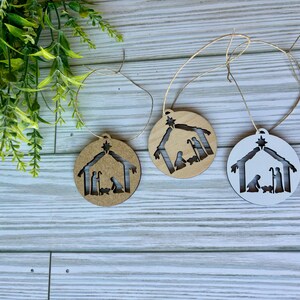 May include: Three wooden Christmas ornaments with a nativity scene cutout. The ornaments are brown, white, and natural wood.