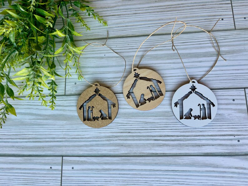 May include: Three wooden Christmas ornaments with a nativity scene cutout. The ornaments are brown, white, and natural wood.