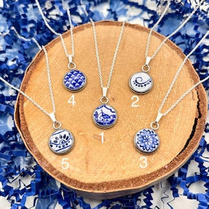 May include: Five silver necklaces with round pendants featuring blue and white designs. The pendants are numbered 1 through 5, showcasing various patterns like floral, geometric, and scenic motifs. The necklaces rest on a wooden surface.