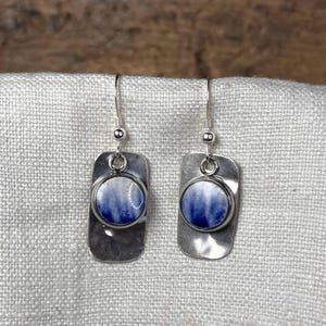 May include: A pair of silver dangle earrings. Each earring features a rectangular, hammered silver base with a circular, blue and white marbled design. The earrings hang from silver fish hook ear wires.