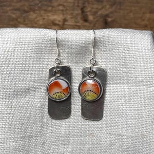 May include: A pair of silver dangle earrings with a rectangular charm and a round, orange, yellow, and white floral design.