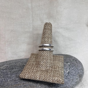 May include: Two silver rings are displayed on a ring holder. The rings are simple bands, one slightly thicker than the other. The ring holder is made of a woven, tan fabric and is placed on a gray stone.