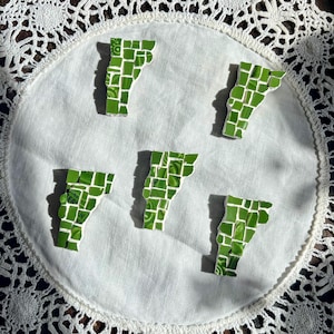May include: Five mosaic Vermont state magnets in shades of green, arranged on a white, lace-edged doily. Each magnet is composed of small, square tiles, creating a textured appearance. The magnets are scattered across the doily.