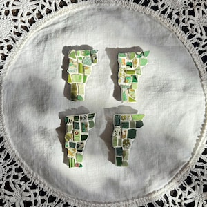 Può includere: Quattro spille a mosaico a forma di stato del Vermont, in varie tonalità di verde, disposte su un panno bianco bordato di pizzo. Ogni spilla è composta da piccoli pezzi irregolari, creando un aspetto strutturato. Le spille variano nei loro toni e motivi verdi.