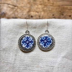 May include: A pair of round earrings with a blue and white floral design. The earrings have a silver-colored border and a simple hook closure. The earrings are displayed on a light-colored fabric.
