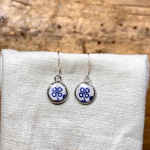 May include: A pair of round, silver-tone dangle earrings. Each earring features a white background with a blue floral design. The earrings are displayed on a white cloth against a wooden background.