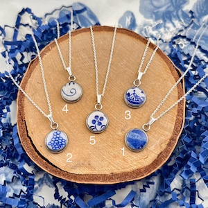May include: Five silver necklaces with round pendants featuring blue and white designs. The pendants are set on a wooden surface with blue paper confetti. The designs include floral patterns and abstract shapes.