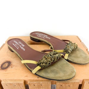 May include: A pair of olive green suede slip-on sandals with a ruffled accent. The sandals have brown insoles and yellow straps. The brand name "DONALD J PLINER" is printed on the insole. The sandals are displayed on a wooden surface.