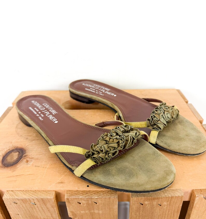 May include: A pair of olive green suede slip-on sandals with a ruffled accent. The sandals have brown insoles and yellow straps. The brand name "DONALD J PLINER" is printed on the insole. The sandals are displayed on a wooden surface.