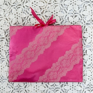 May include: A bright pink satin pouch with diagonal lace trim and a matching ribbon tie. The lace trim is a lighter shade of pink, creating a delicate contrast. The pouch is rectangular in shape.