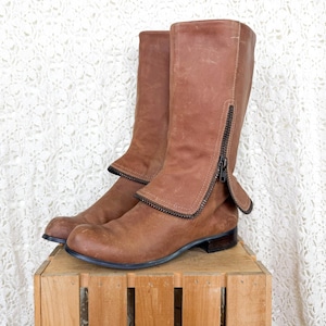 May include: A pair of mid-calf boots in a medium brown leather finish. The boots feature a rounded toe, a low block heel, and a distinctive fold-over cuff detail on the outer side secured by a dark metal zipper that runs vertically along the shaft.