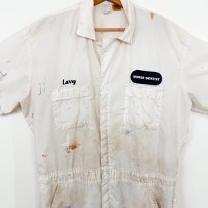 May include: A vintage white jumpsuit with short sleeves, featuring two chest pockets and a front zipper. The jumpsuit has paint stains and the name "Larry" embroidered on the chest. A patch that says "HORSE DENTIST" is also visible.
