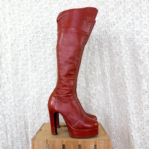 May include: A knee-high boot in a deep red color, crafted with a smooth finish. The boot features a high block heel and a thick platform sole, with a structured shaft that reaches just below the knee and a clean, minimalist design throughout.