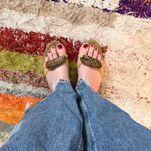 May include: A top-down view of a person's feet wearing olive green sandals with floral accents. The person is wearing blue denim jeans. The background is a colorful, textured rug with red, green, orange, and white stripes.