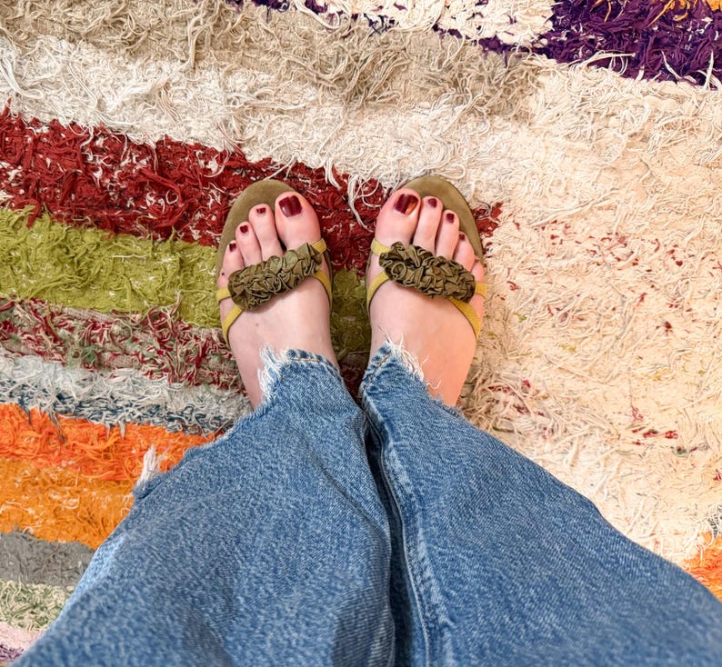 May include: A top-down view of a person's feet wearing olive green sandals with floral accents. The person is wearing blue denim jeans. The background is a colorful, textured rug with red, green, orange, and white stripes.