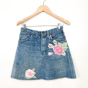 May include: A denim skirt with a light blue wash, adorned with pink rose appliqués and green leaves. The skirt has a frayed hem and is displayed on a wooden hanger.