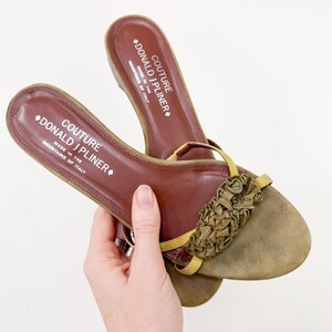 May include: Brown leather sandals with a decorative ruffle detail on the toe. The sandals have a thong strap and a cushioned footbed. The brand name "DONALD J PLINER" is visible on the insole. Made in Italy.
