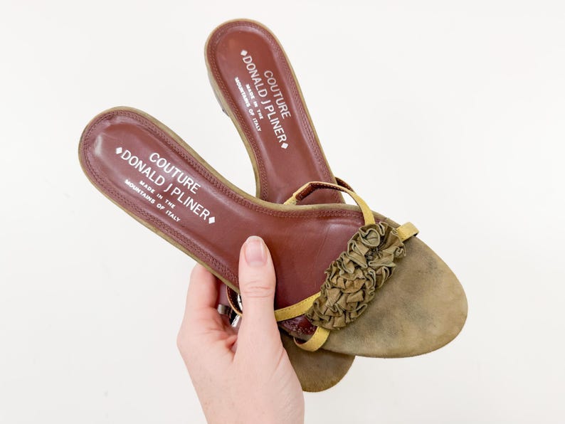 May include: Brown leather sandals with a decorative ruffle detail on the toe. The sandals have a thong strap and a cushioned footbed. The brand name "DONALD J PLINER" is visible on the insole. Made in Italy.