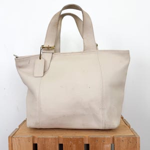 May include: Cream-colored leather tote bag with two handles and a small tag. The bag has a rectangular shape with a zipper closure and a gold-colored metal clasp. The bag is sitting on a wooden crate.