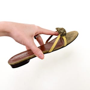 May include: A single olive green suede sandal with a yellow strap and a decorative floral accent. The sandal has a brown sole and a low heel. The brand name "DONALD J PLINER" is visible on the sole.