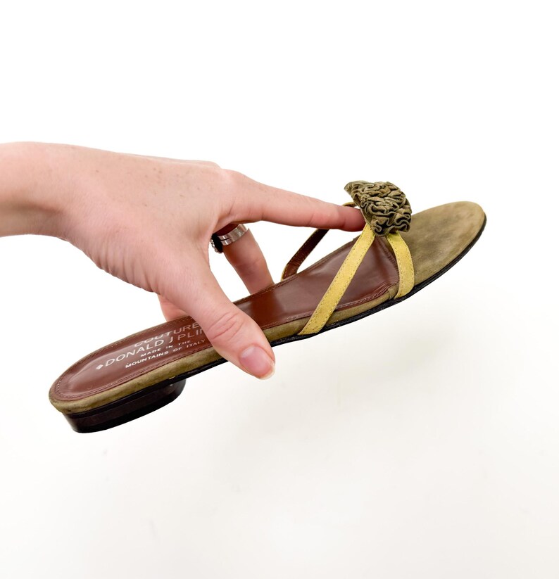 May include: A single olive green suede sandal with a yellow strap and a decorative floral accent. The sandal has a brown sole and a low heel. The brand name "DONALD J PLINER" is visible on the sole.